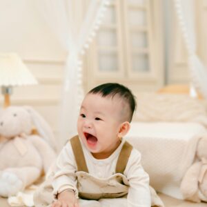 Charming Asian baby laughing indoors surrounded by soft toys in a cozy room.